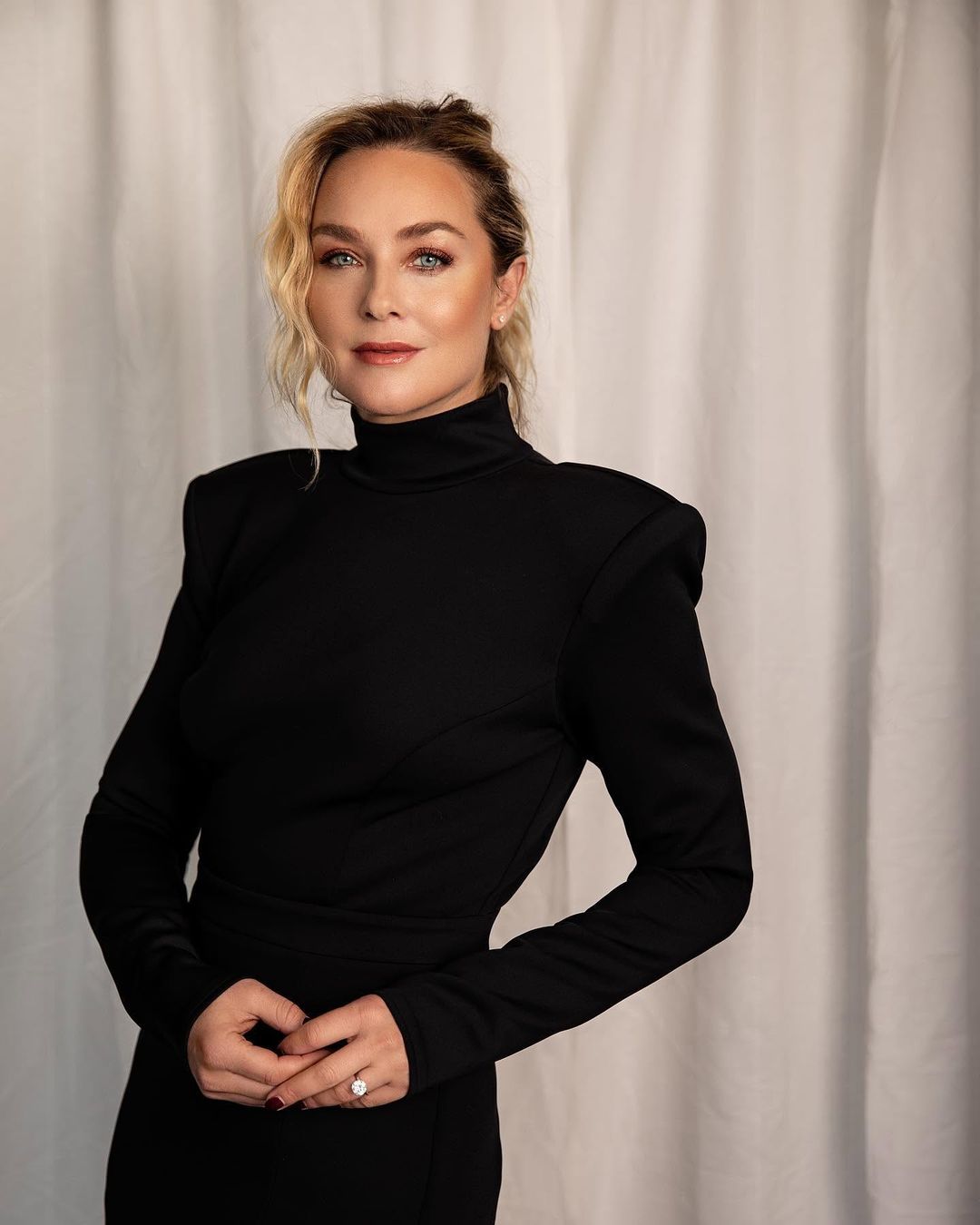 Elisabeth Röhm in a black strong-shouldered turtleneck dress against a white curtain for Jejune Magazine, styled by Quentin Fears