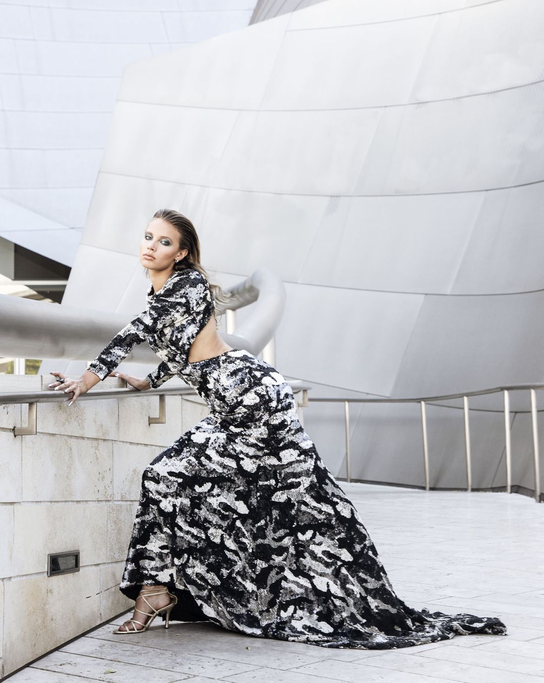 Industry Magazine editorial: a black and silver camouflage sequin gown with an open back and a long train, leaning on a rail before the steel facade of Walt Disney Concert Hall, styled by Quentin Fears