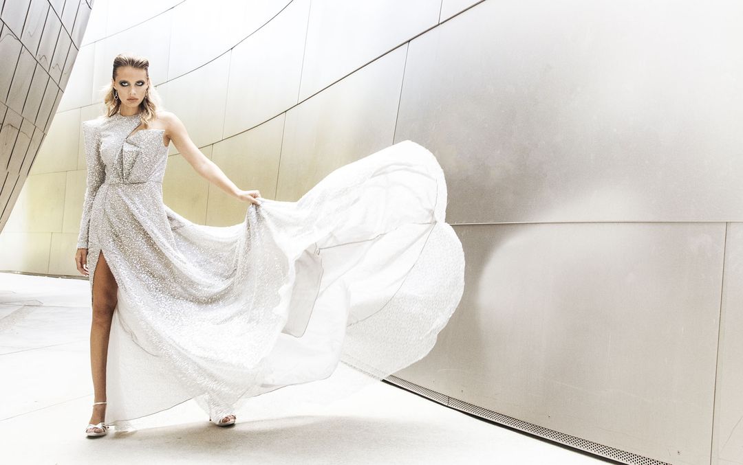 Industry Magazine editorial: a silver sequin one-shoulder gown with a sharp shoulder and a sweeping chiffon train held out against the curved steel walls of Walt Disney Concert Hall, styled by Quentin Fears
