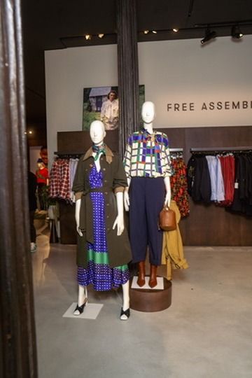 Two mannequins in the Free Assembly section: a green trench over a printed dress, and a graphic-print shirt with navy trousers